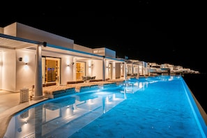 A modern infinity pool with a clear edge, adjacent to a building with a well-lit entrance and outdoor seating area.