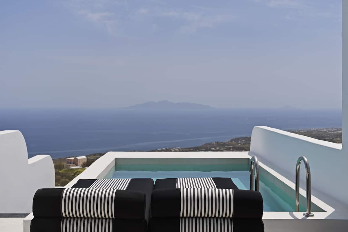 Ein Infinity-Pool auf dem Dach mit schwarz-weiß gestreiften Liegestühlen mit Blick auf eine Küstenlandschaft und entfernte Berge.