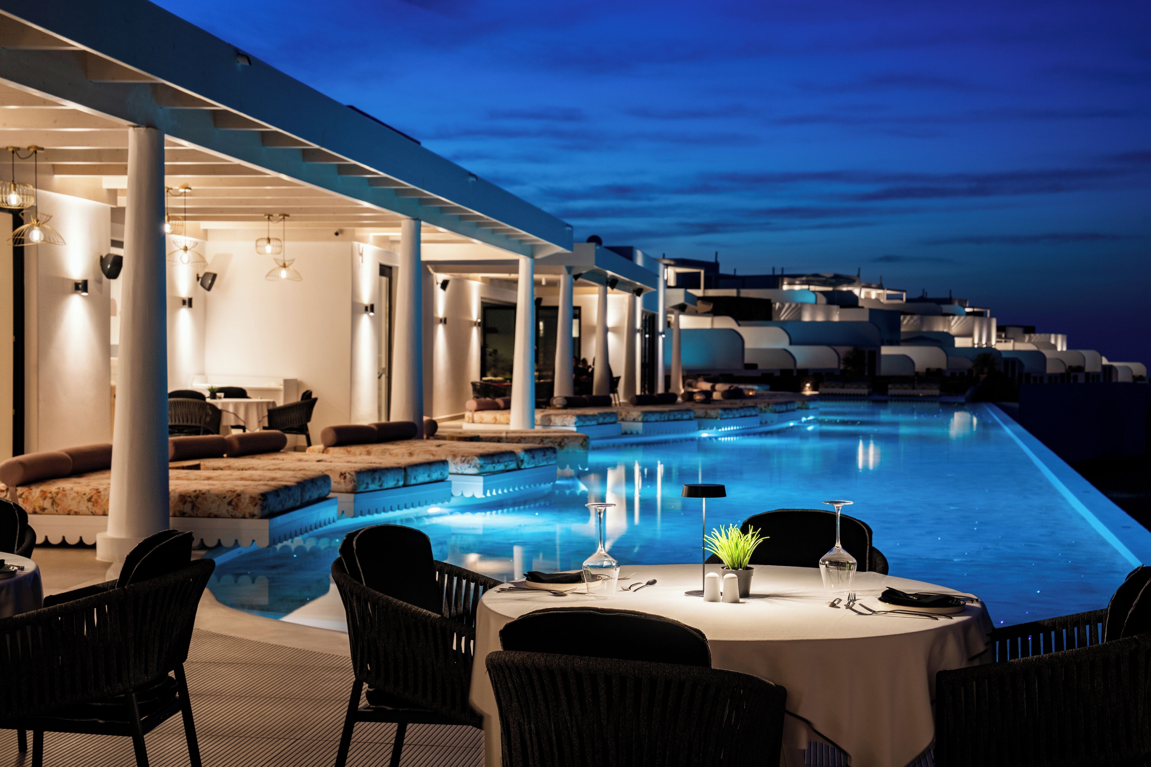 An infinity pool with a dining area, outdoor seating, and a view of the sea at dusk.