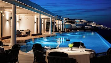 An infinity pool with a dining area, outdoor seating, and a view of the sea at dusk.