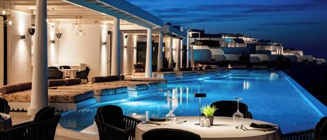 An infinity pool with a dining area, outdoor seating, and a view of the sea at dusk.