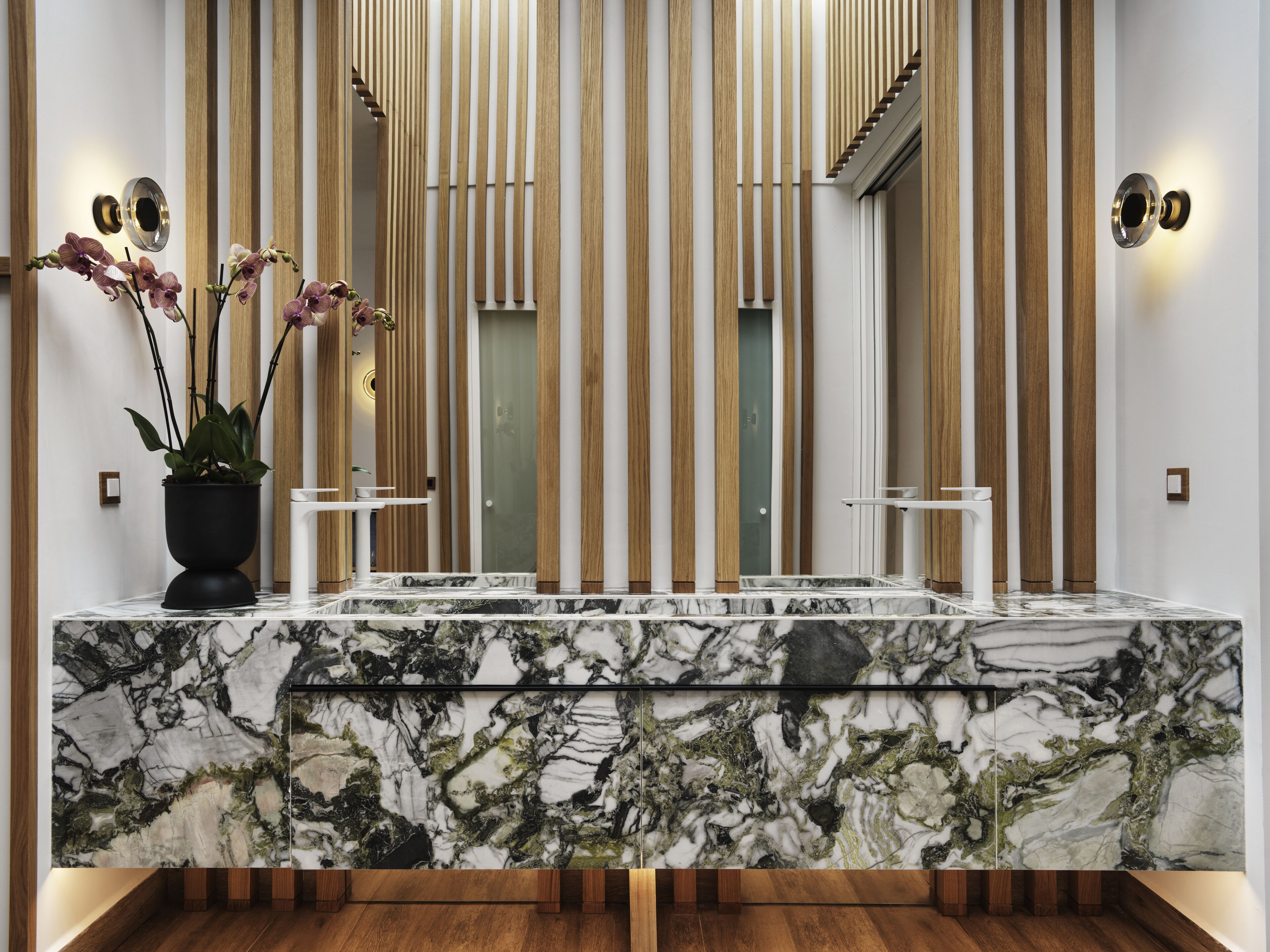 A modern bathroom with a marble countertop, wooden paneling, and a potted orchid plant.