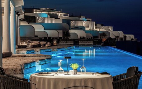 A dining area with a table set for two, overlooking a pool with a modern architectural backdrop.