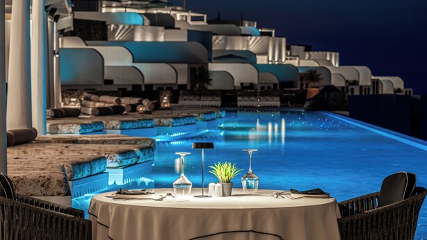 A dining area with a table set for two, overlooking a pool with a modern architectural backdrop. - Cresanto Luxury Suites  (Santorini)