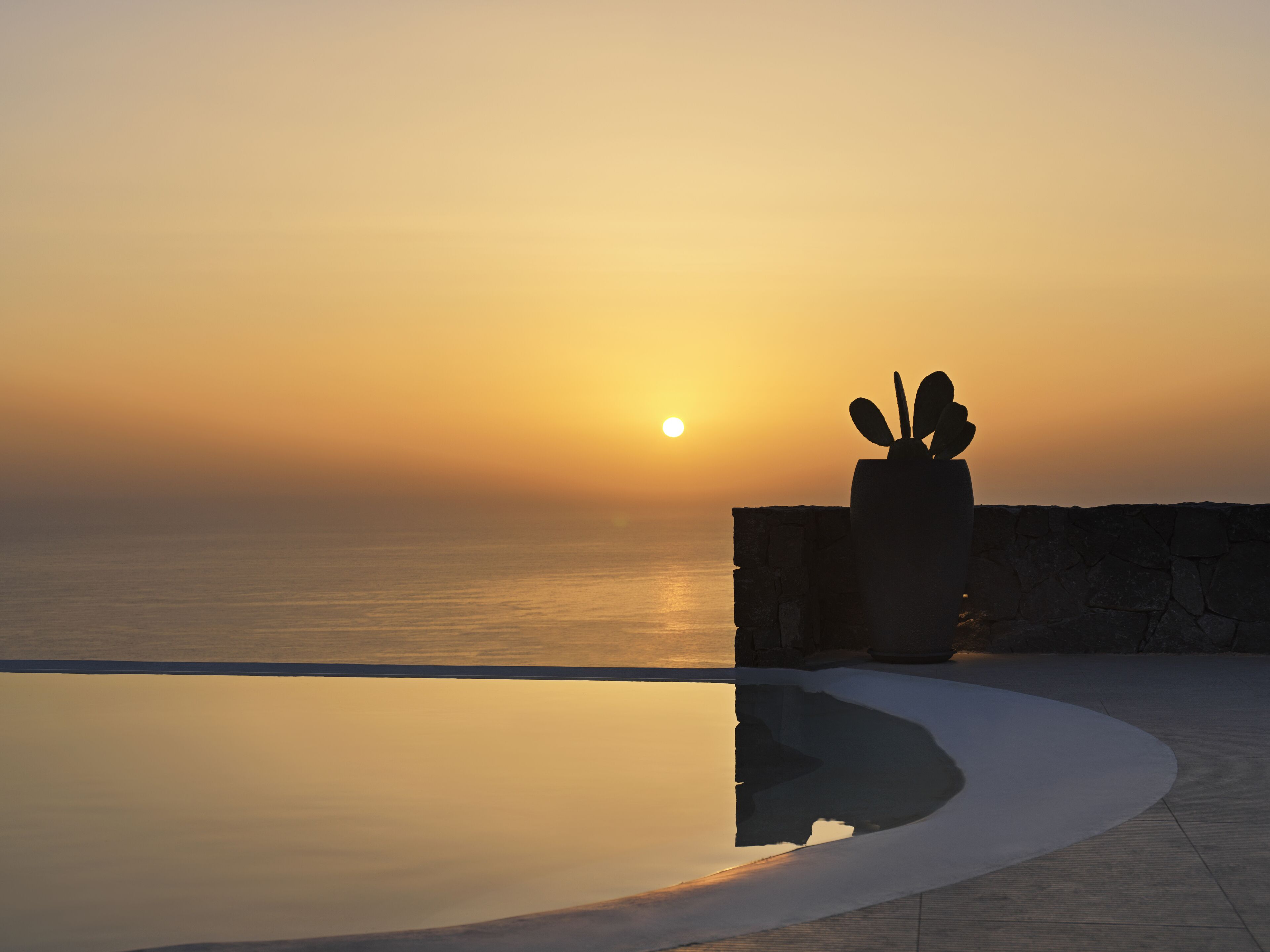 A sunset over a calm sea with a stone wall and a cactus sculpture.