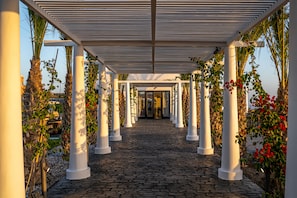 A covered walkway with white columns and cobblestone flooring, leading to a building entrance.