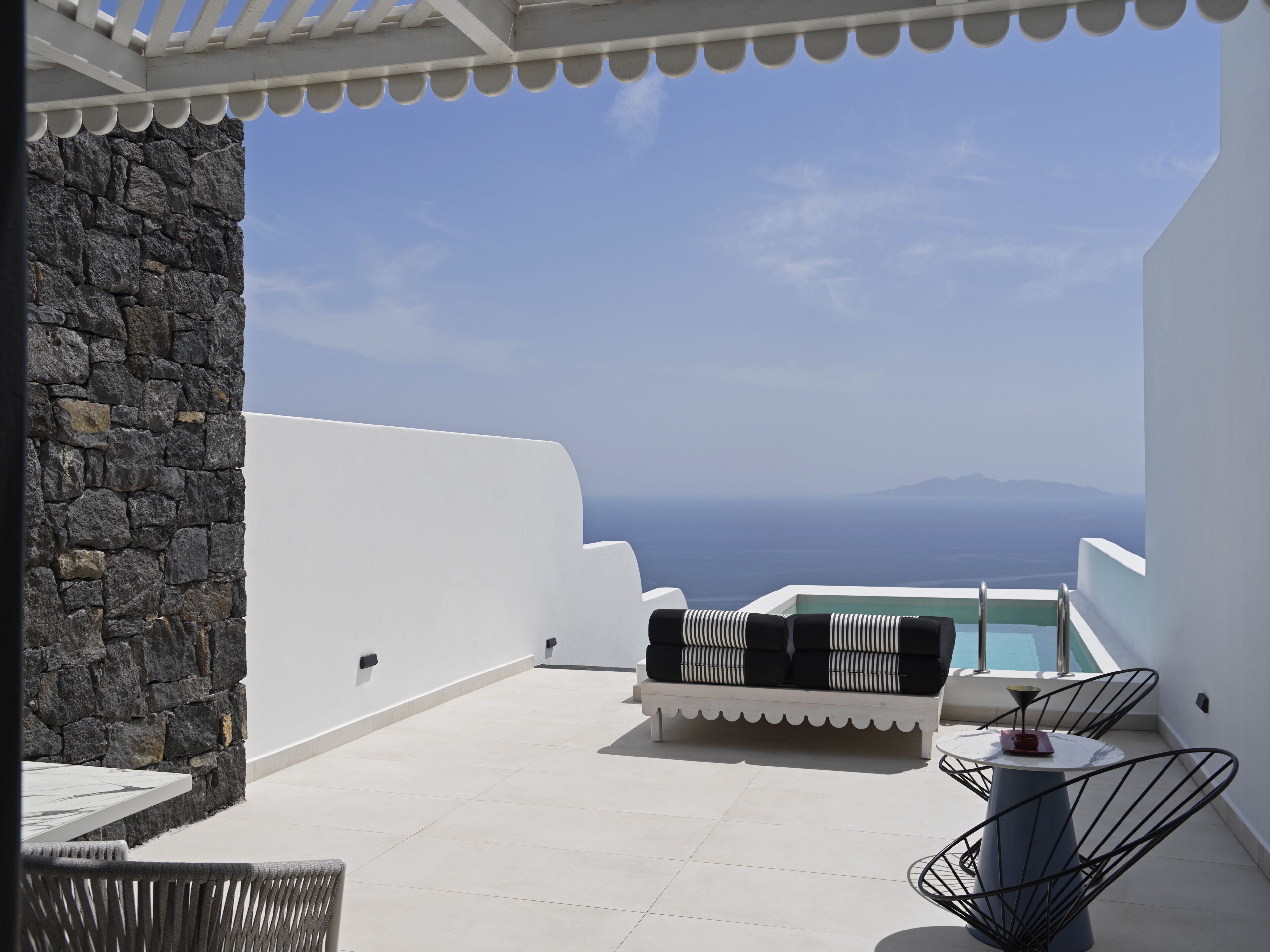 A terrace with a stone wall, white walls, a white sofa with striped cushions, a round table, and a view of the sea and distant mountains.