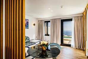 A modern hotel room with a glass table, a sofa, a chair, and a view of the sea through a large window.