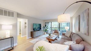 A modern living room with a sofa, coffee table, and TV. There is a balcony with a view of trees. - Stunning, coastal chic two bedroom, two bathroom villa in Palmetto Dunes!! (Hilton Head)