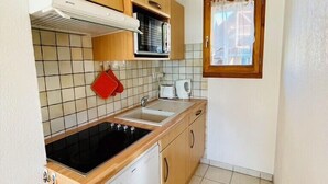 A compact kitchen with wooden cabinets, a stove, a sink, and a window with a curtain. - Nice Cosy Apt With Balcony In Briançon (Briançon)
