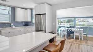 A modern kitchen with white cabinets, a stainless steel refrigerator, and a marble island. There is a dining area with white chairs and a table. - 🌊Updated Vintage Beach House-Private Pool, Steps to Beach-Blue Lizard🦎 (Fort Myers Beach)