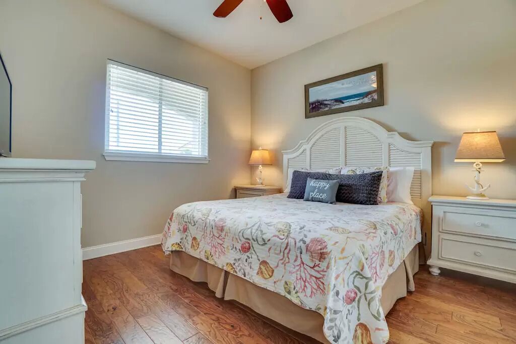 A bedroom with a bed, two bedside tables, a ceiling fan, a window with blinds, and a painting on the wall.