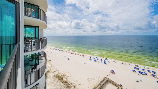 A beachfront hotel with multiple balconies overlooking the ocean, a sandy beach with umbrellas, and a clear view of the sea.