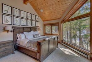 Een slaapkamer met een groot bed, een houten hoofdbord, meerdere ingelijste botanische prints aan de muur en uitzicht op bomen door grote ramen.
