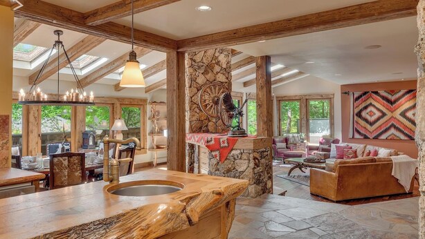 Un intérieur spacieux avec une cheminée en pierre, des poutres apparentes, un coin repas et un salon doté d’une grande fenêtre.