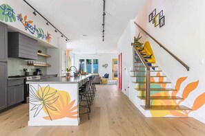 A modern kitchen with a central island, a staircase with colorful railings, and a living area with a sofa and chairs.