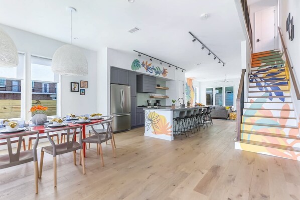 A modern kitchen with a central island, a dining area with a red table, and a staircase with colorful steps.