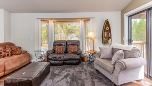 A living room with a brown leather sofa, a beige sofa, and a brown leather recliner. There is a small table with a lamp, a bookshelf, and a window with blinds. - BLUE01 - Spacious Home, AC, SHARC Passes, Hot Tub (Sunriver)