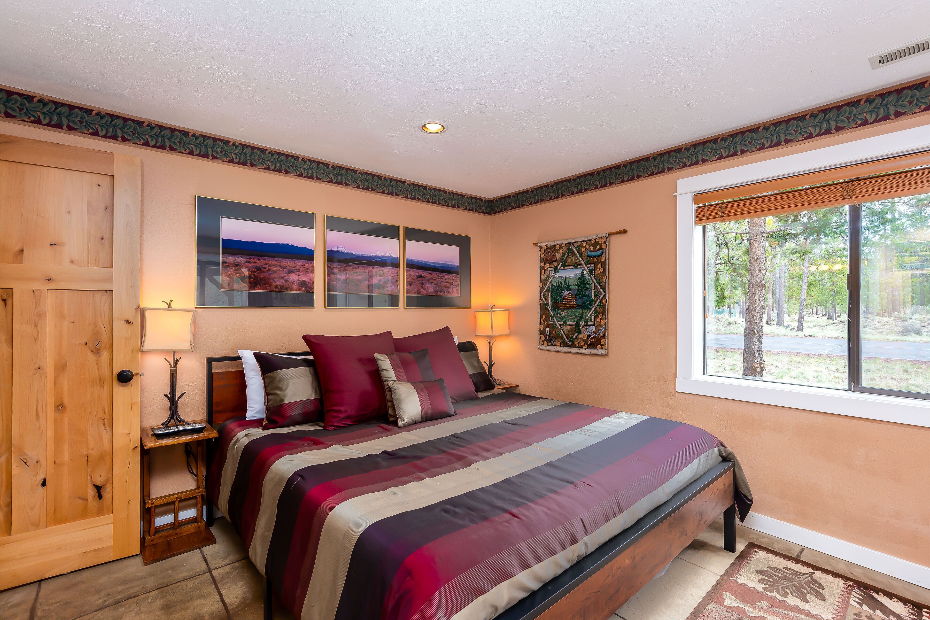 A bedroom with a bed, a wooden door, a window with a view, and a wall with three framed pictures.