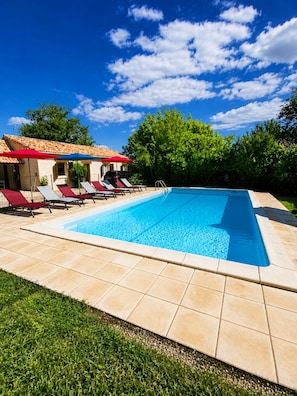 A swimming pool with lounge chairs and umbrellas, surrounded by a tiled deck and greenery.