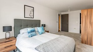 A modern bedroom with a large bed, a grey upholstered headboard, bedside tables with lamps, and a framed artwork on the wall. - Stay Zeni – Contemporary 2 BR Flat in York, Ouse Court (York)