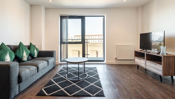 A modern living room with a grey sofa, a small round table, a TV on a wooden stand, and a large window with a view of buildings. - Stay Zeni – Contemporary 2 BR Flat in York, Ouse Court (York)