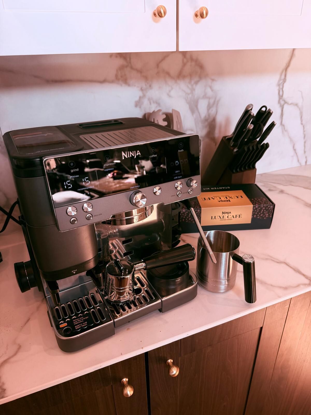 Une machine à expresso Ninja Luxe Café, posée sur un comptoir en marbre, avec un moulin à café et une boîte de capsules Ninja Luxe Café.