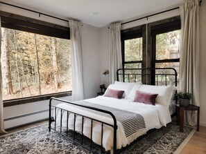 A bedroom with a bed, two bedside tables, a window with curtains, and a view of trees.