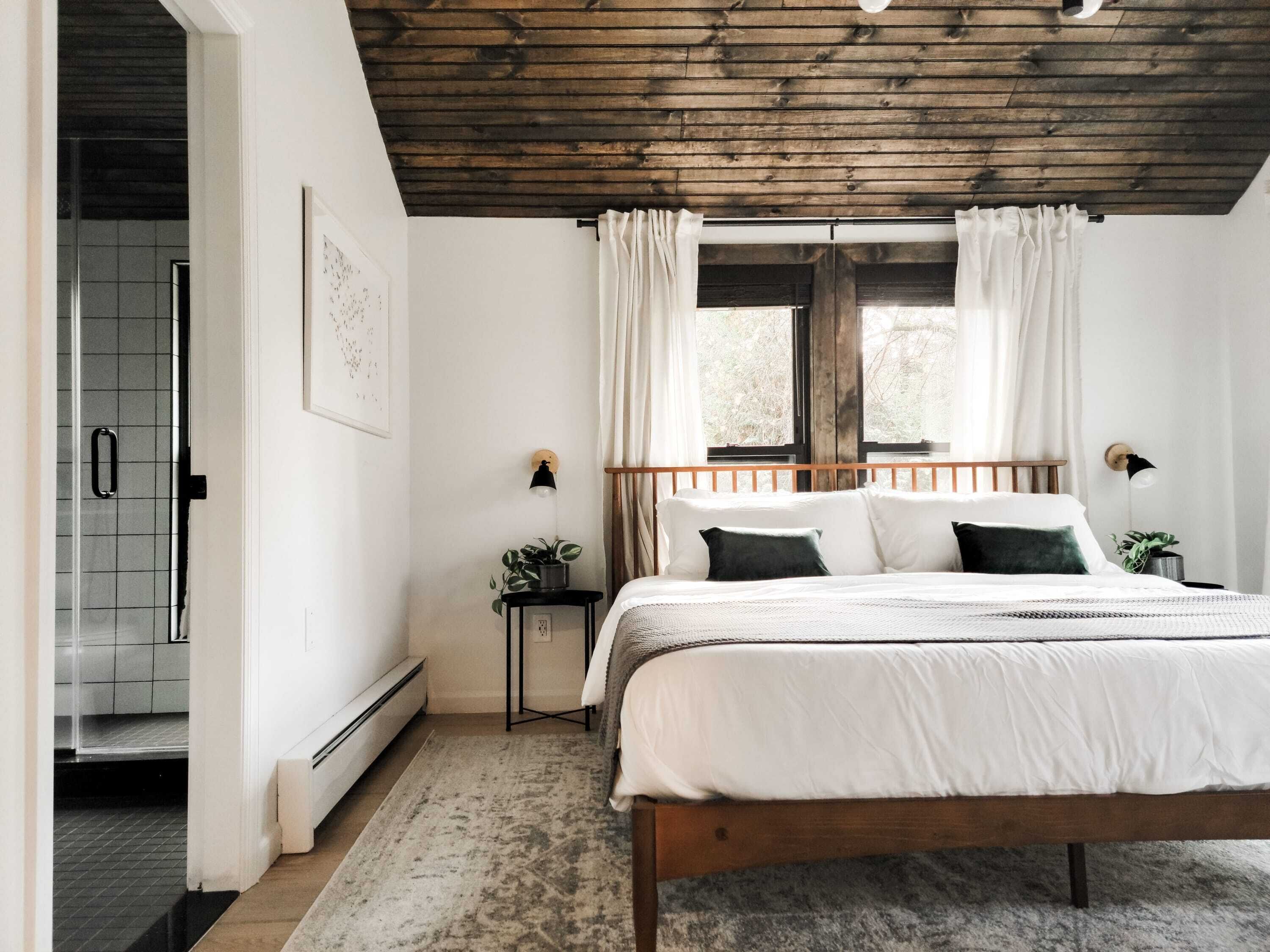 Een slaapkamer met een houten bed, witte beddengoed, een klein nachtkastje met een lamp en een raam met gordijnen.
