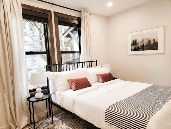 Une chambre à coucher avec un lit, une table de chevet avec une lampe, une fenêtre avec des rideaux et un tableau encadré au mur.