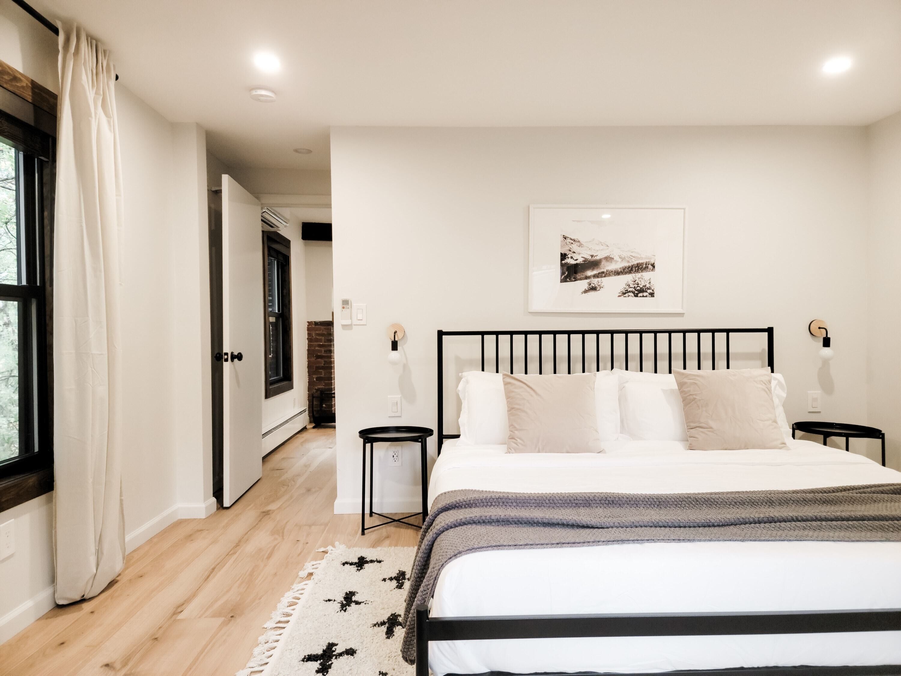 A bedroom with a large bed, a black headboard, and a wooden floor. There is a window with curtains, a wall-mounted light, and a framed picture above the bed.