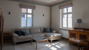En stue med en sofa, skammel og et lille bord. To vinduer med gardiner, en radiator og en lampe på et træskab.
