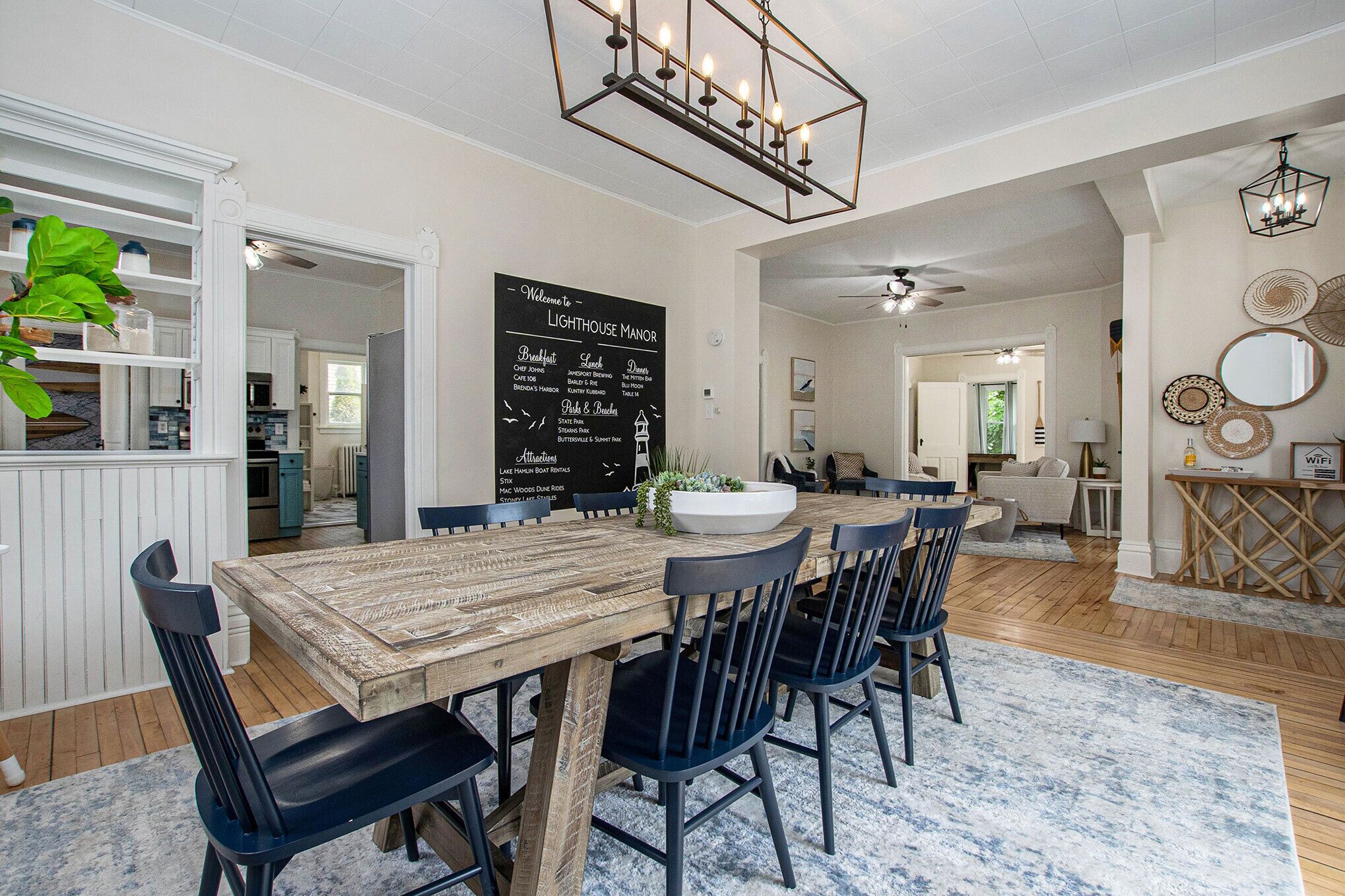 Un espace repas avec une table en bois et des chaises noires, un tableau noir sur lequel est écrit un message de bienvenue, et un ventilateur de plafond.