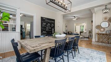 Un espace repas avec une table en bois et des chaises noires, un tableau noir sur lequel est écrit un message de bienvenue, et un ventilateur de plafond.
