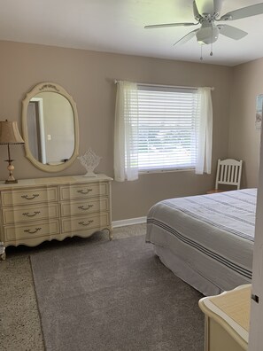Un dormitorio con cama, cómoda, espejo, ventana con persianas, ventilador de techo y una silla.