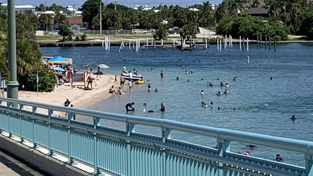Ein Uferbereich mit Strand, badenden Personen und einer Brücke mit Geländern.