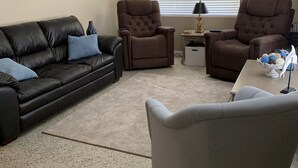 Ein Wohnzimmer mit einer schwarzen Ledersofa, zwei braunen Sesseln, einem grauen Sessel, einem Deckenventilator, einem Fenster mit Jalousien und einer Wand mit drei gerahmten Bildern.