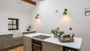 A kitchen with a central island, wooden beams, pendant lighting, a window with blinds, and a built-in oven. - Yr Hen Oriel (Caernarfon)