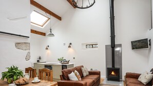 A cozy living room with a fireplace, a dining area with a wooden table, and a kitchen with a window. - Yr Hen Oriel (Caernarfon)