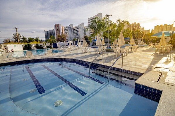 Piscina con sillas de descanso y sombrillas, rodeada de edificios.