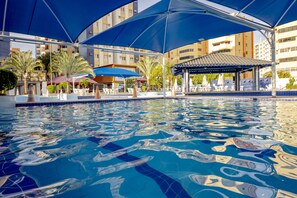 Una piscina con bordes de azulejos azules, rodeada de sombrillas y sillas de descanso, con edificios al fondo.