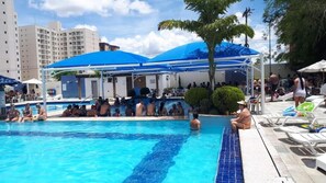 Piscina com pessoas ao redor, área coberta e edifícios ao fundo.