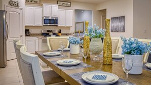 A dining area with a long table set for six, white cabinets, a kitchen in the background, and decorative vases with blue flowers.
