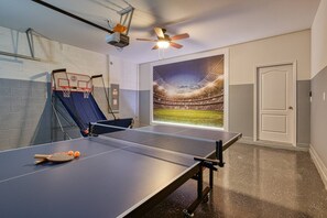 Una mesa de ping pong con red, un proyector montado en la pared y un ventilador de techo en una habitación con un gran mural de un estadio deportivo.