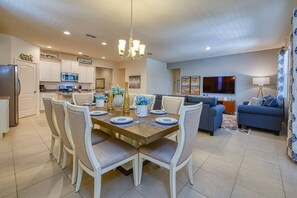 Una cocina moderna con armarios blancos, un comedor con mesa y sillas de madera, y una sala de estar con sofá azul y televisor de pantalla plana.