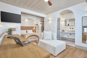 A modern hotel room with a wooden desk, a laptop, a comfortable sofa, a bed, a TV mounted on the wall, and a bathroom with a mirror and shelves. - GrandeMar Suites (Astypalaia)