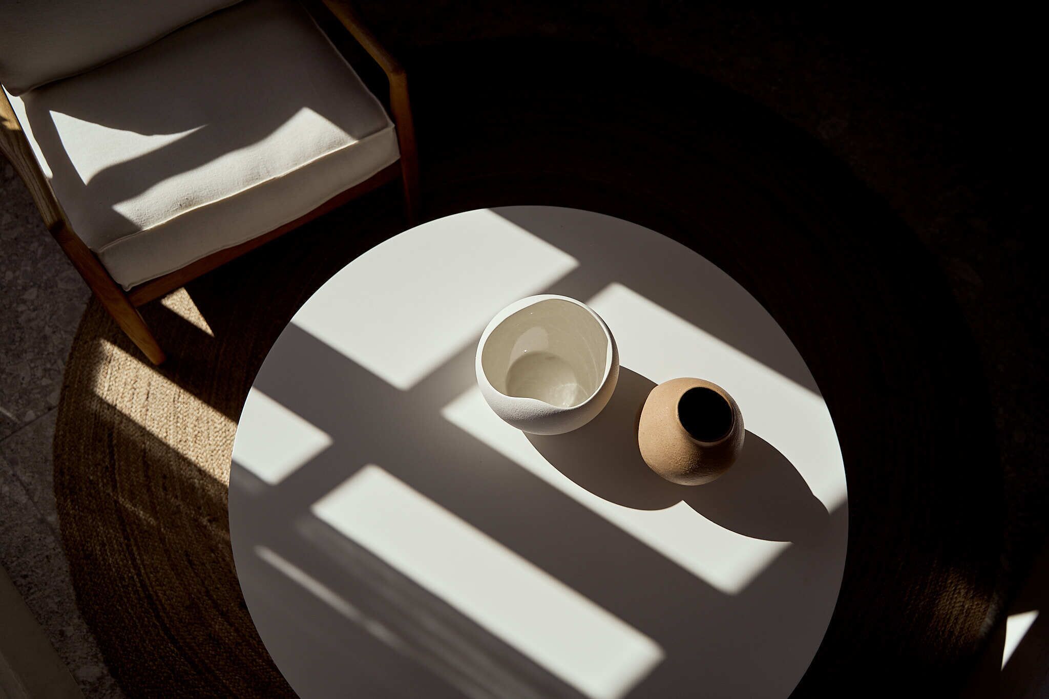 Une table ronde avec un bol blanc et un petit récipient, une chaise en bois avec un coussin, et la lumière du soleil projetant des ombres sur le sol.