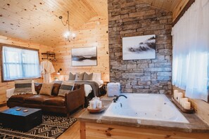 Interior acogedor de una cabaña con techo de madera, pared de piedra, cama con muchos cojines, sofá de cuero, una mesita y una bañera con velas.