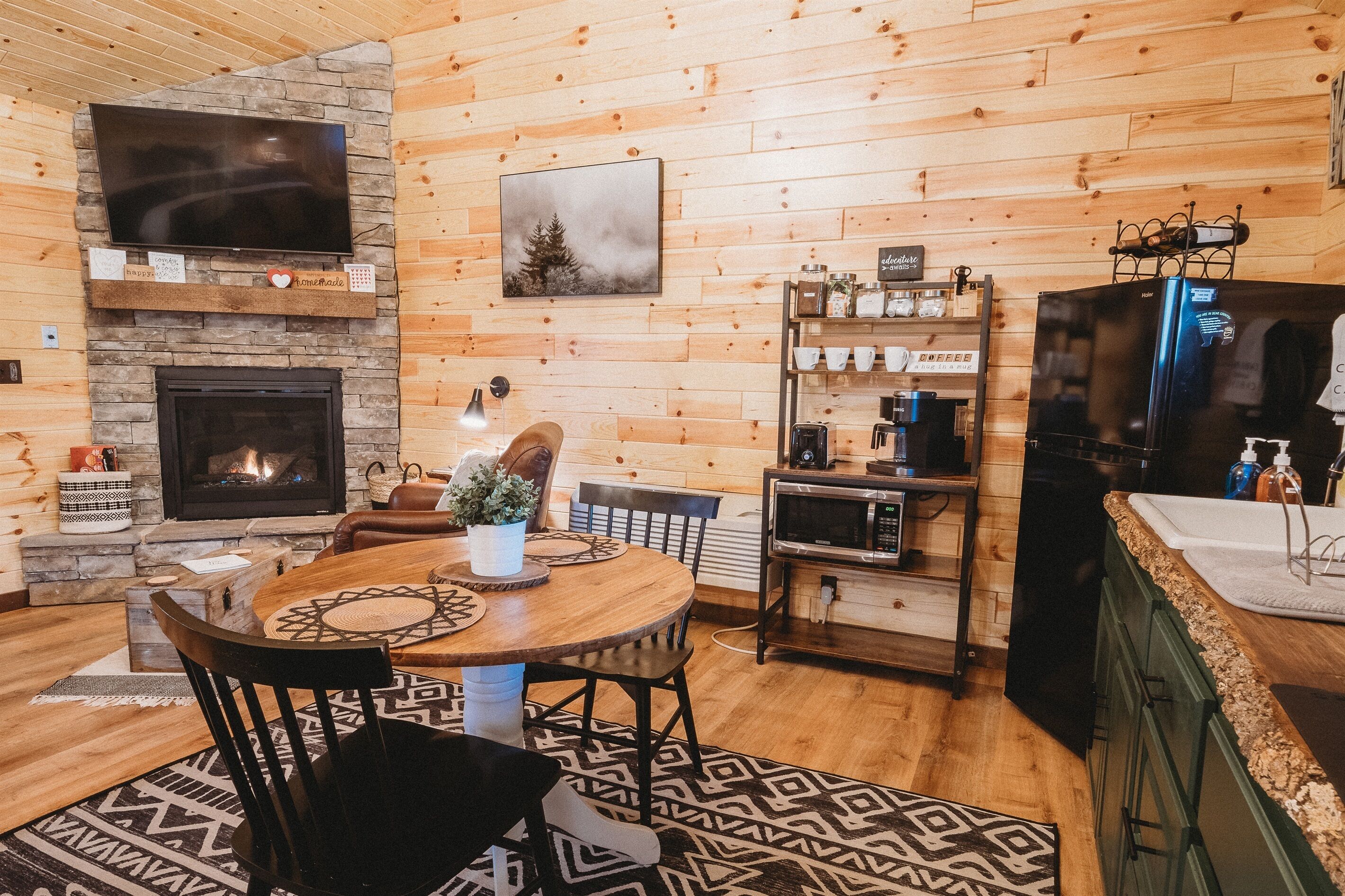 Un intérieur de chalet chaleureux avec une cheminée en pierre, des murs en bois, un coin repas avec une table ronde et une cuisine équipée d’un réfrigérateur et d’une machine à café.