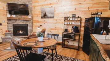 Un intérieur de chalet chaleureux avec une cheminée en pierre, des murs en bois, un coin repas avec une table ronde et une cuisine équipée d’un réfrigérateur et d’une machine à café.